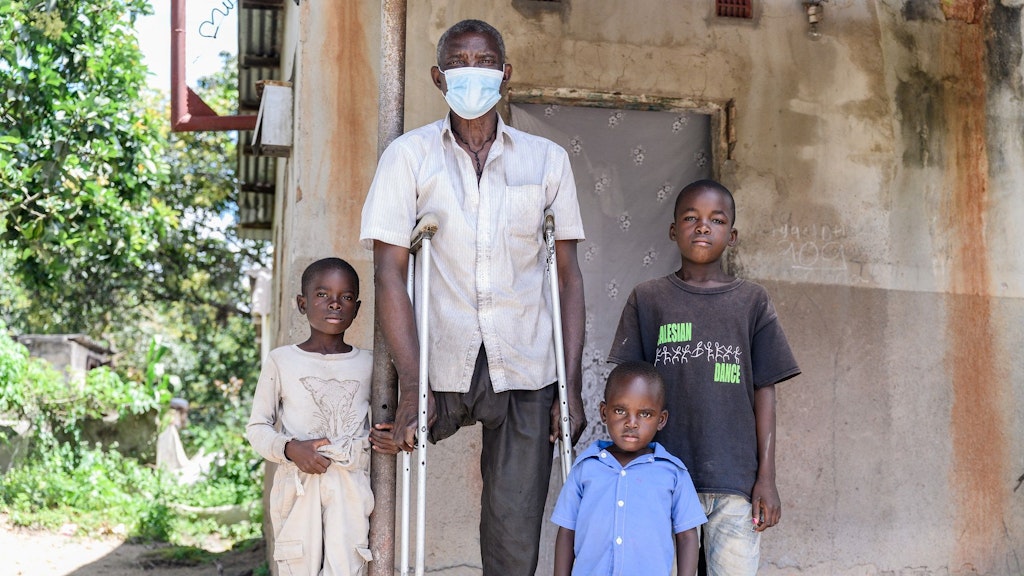 An older man with one leg, supported by crutches, standing in front of a house surrounded by three children of different ages.
