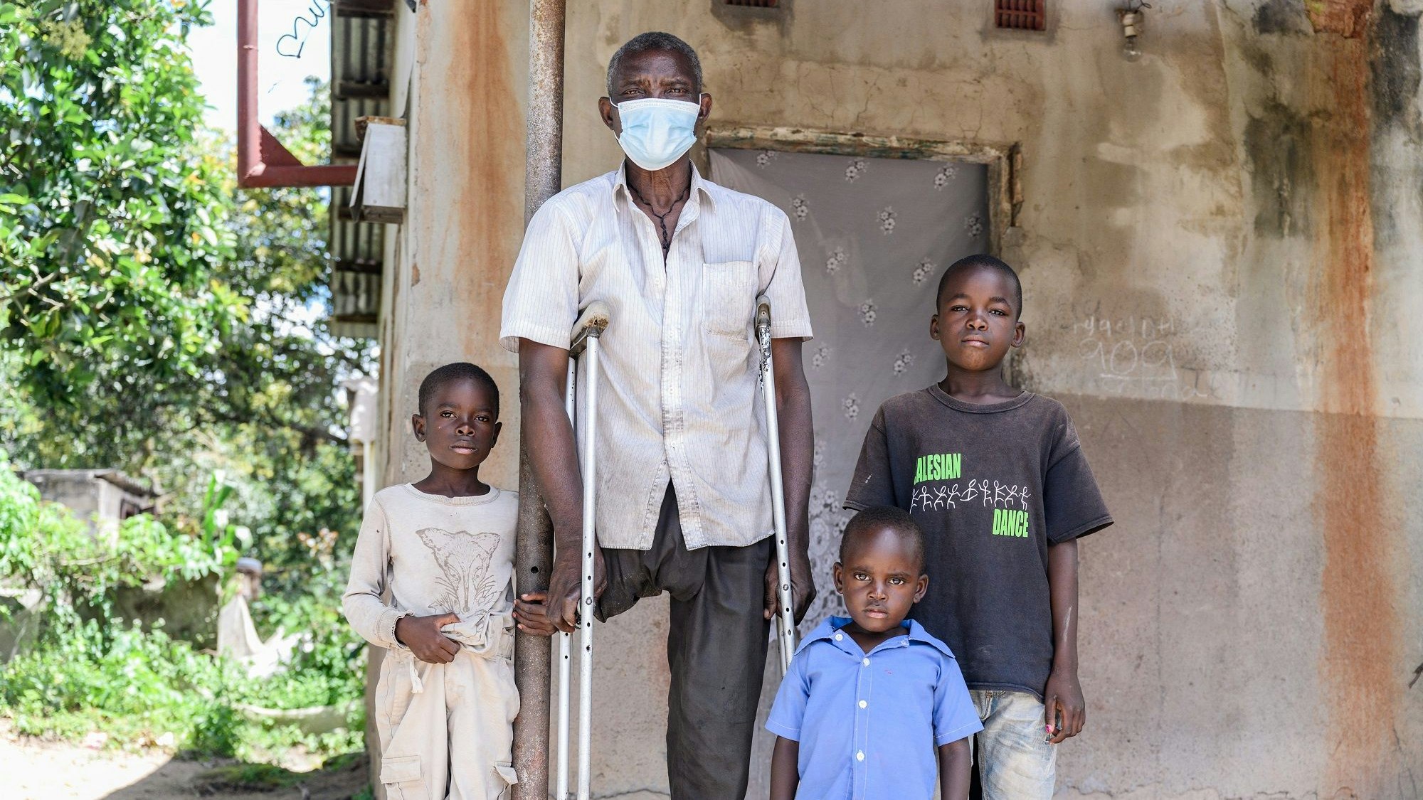 An older man with one leg, supported by crutches, standing in front of a house surrounded by three children of different ages. 