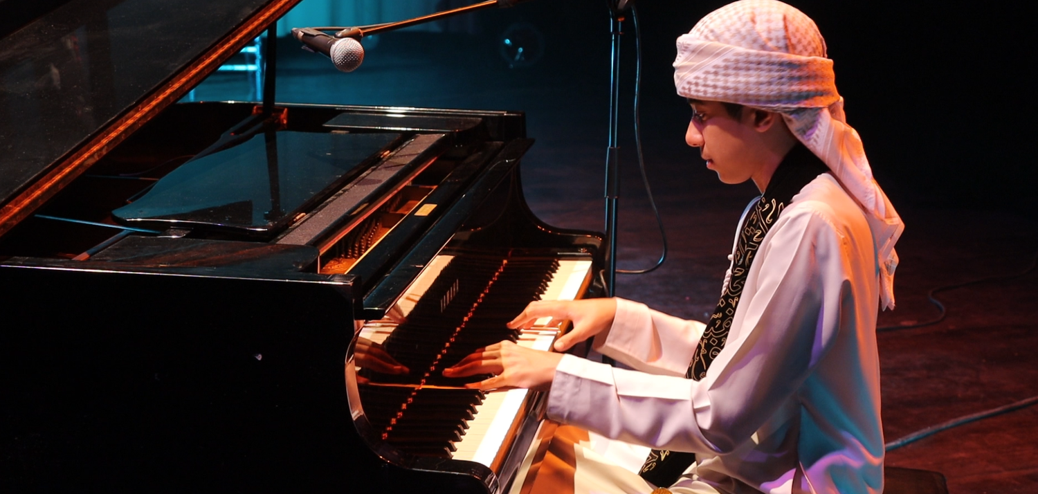 A young man dressed in traditional costume plays the piano on a dimly lit stage.